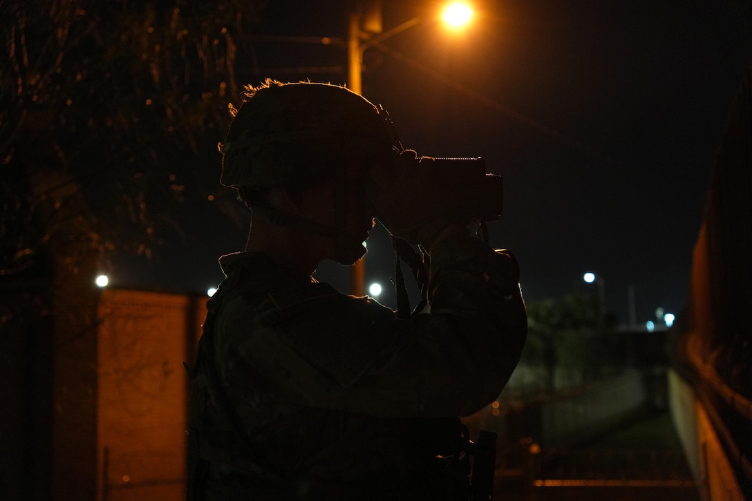 A soldier uses binoculars while standing in the dark near a street light illuminated by a yellowish, orange light.