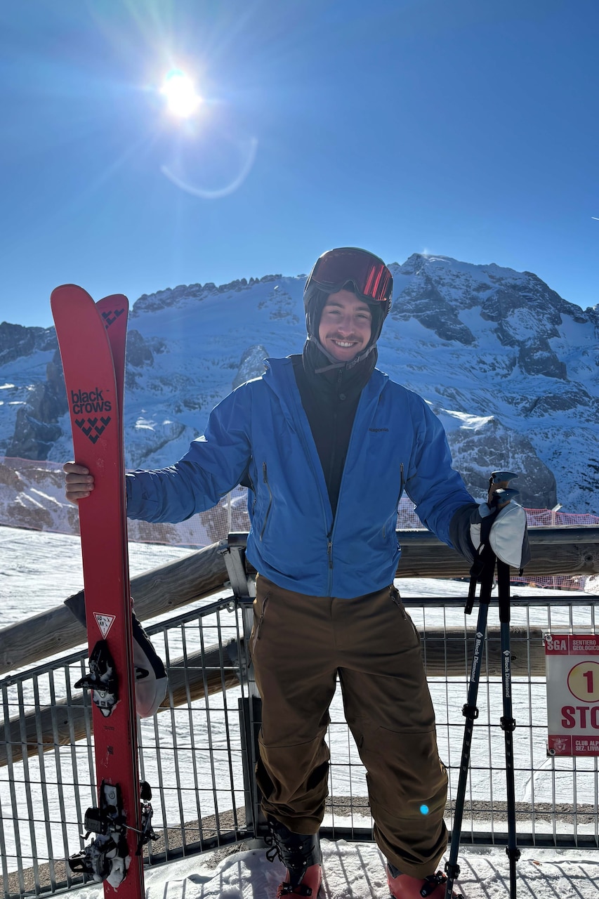 An airman poses with skies and ski poles on top of a snowy mountain during a sunny day.