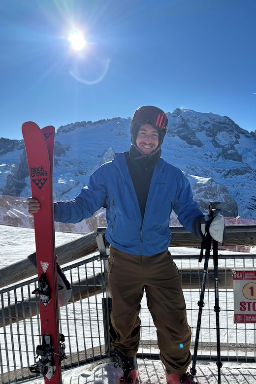 An airman poses with skies and ski poles on top of a snowy mountain during a sunny day.