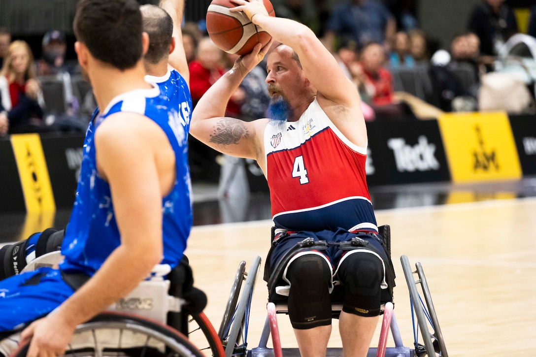 A veteran with a blue beard using a wheelchair passes a ball during a basketball game as spectators watch from bleachers in a blurred background.