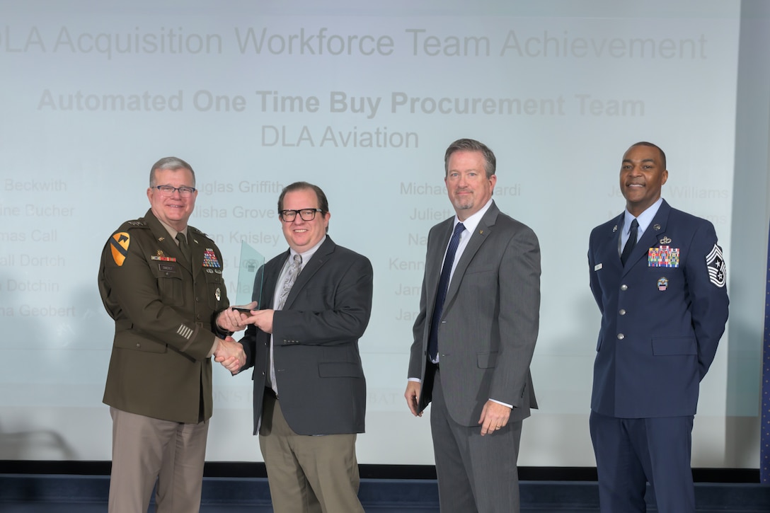 A man receives an award from a military officer. Two other leaders are pictured.