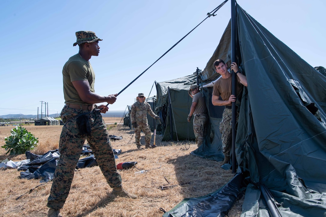 Three soldiers pitch a tent on a field during a clear day while another soldier observes.