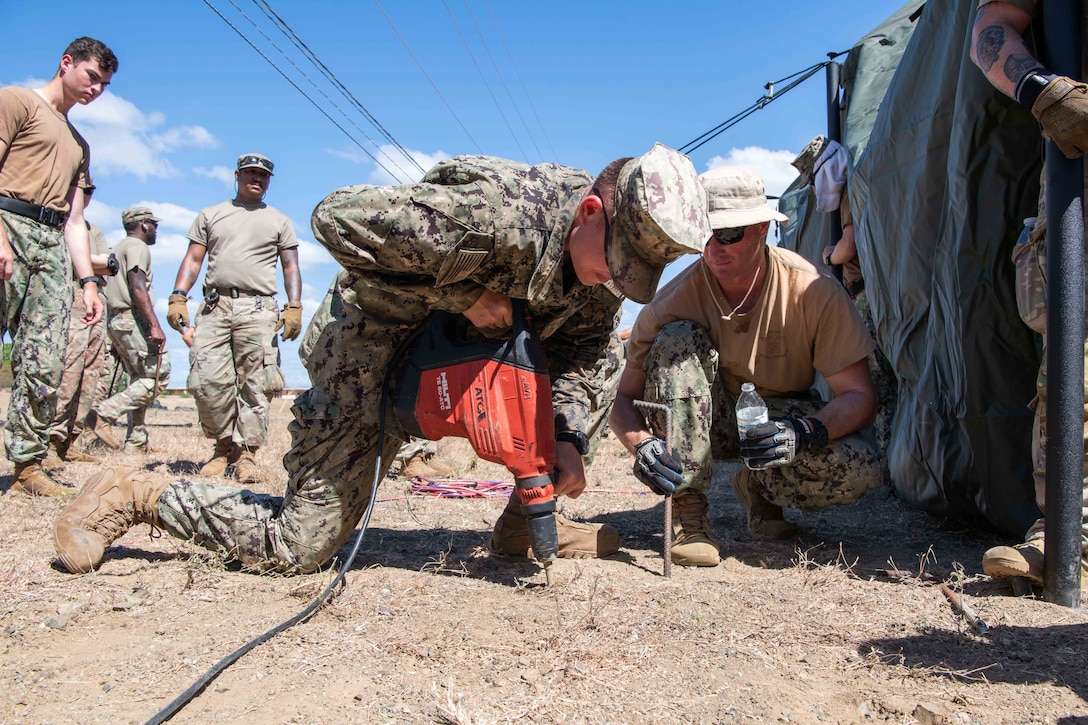 A soldier drills a hole in the ground while another soldier holds a tent pitch and water bottle during the day.