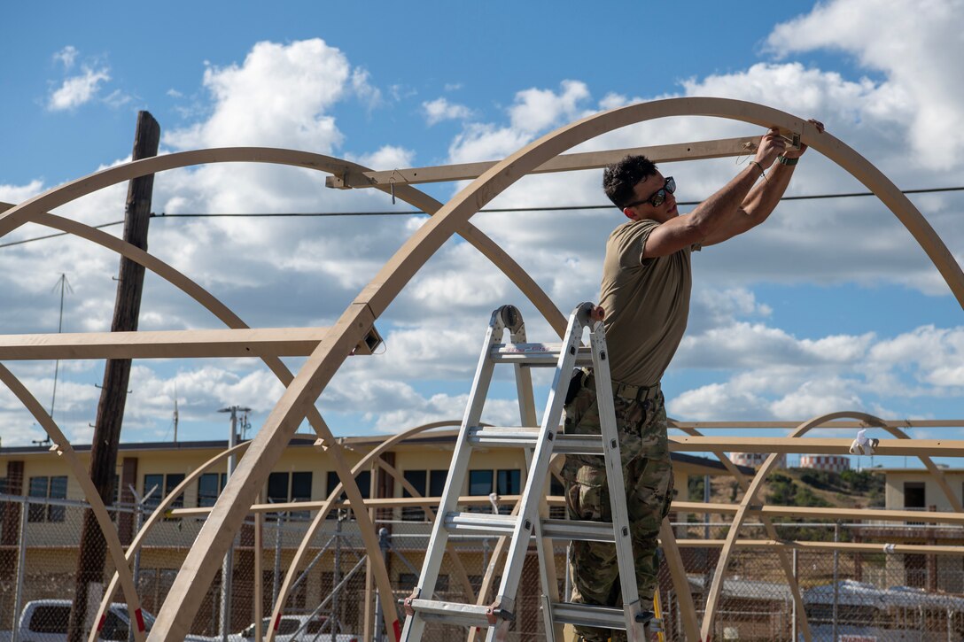 An airman standing on a ladder works on the wooden scaffolding of a tent under partly cloudy skies.