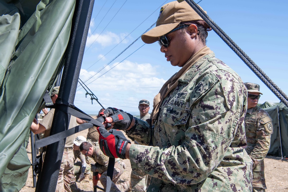A soldier ties a tent to a tent pole during the day at a military base.