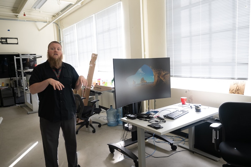 A man uses his hands to talk in a room. A large computer desk is beside him.