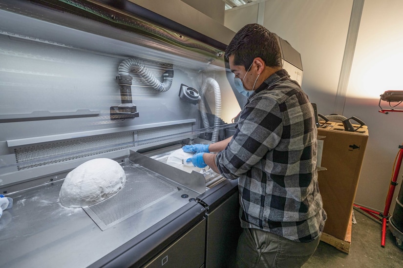 A man in gloves works to uncover a piece of 3D-printed equipment from a pile of powder.