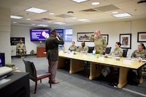 An Army soldier is standing and saluting toward another soldier who is behind a conference room table where four other soldiers are seated.