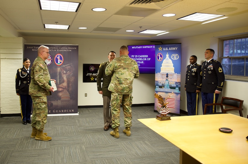 Several Army soldiers in various uniforms are standing in different places around a conference room while one Soldier is being recognized by a general officer.