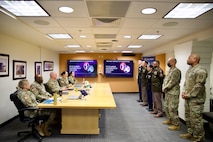 Four Army Soldiers in various uniforms as standing in front of a table where other Soldiers are seated. There are two TV monitors on the wall in the background.