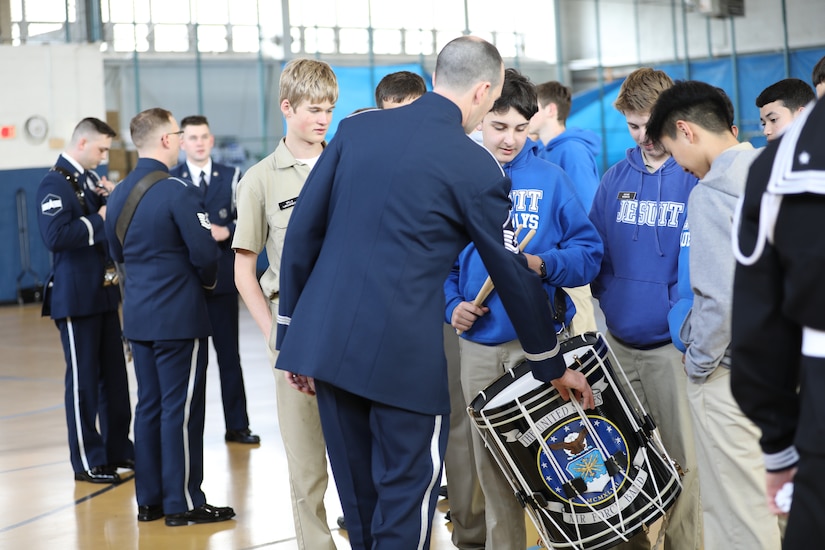 An Air Force Airman is leaning over to show high school students a drum. They are in a gymnasium.