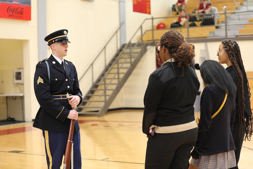 An Army soldier dressed in ceremonial uniform and holding a rifle is talking with students in a gymnasium.