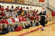 A Navy Sailor is gesturing while talking with a large crowd of high school students, who are sitting on bleachers in a gymnasium.