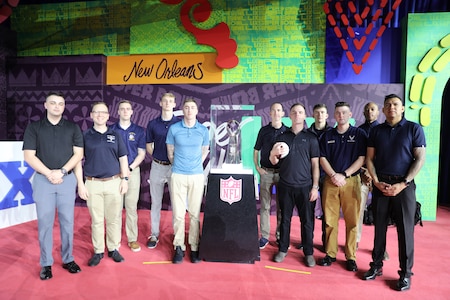 Service members dressed in civilian clothes are posing with the Lombardi Trophy, which is inside a glass case.