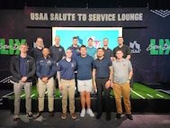 Service members dressed in civilian attire pose with a football player at an NFL event. Above them a sign reads "USAA Salute to Service Lounge."