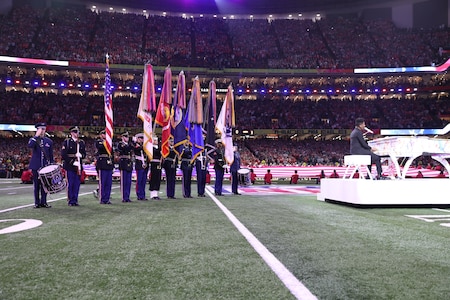 Joint service members are presenting the colors on a football field. There is a musician at a white piano on the right.