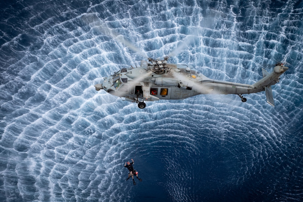 A helicopter hovers over the ocean while a sailor hangs from the helicopter below it.