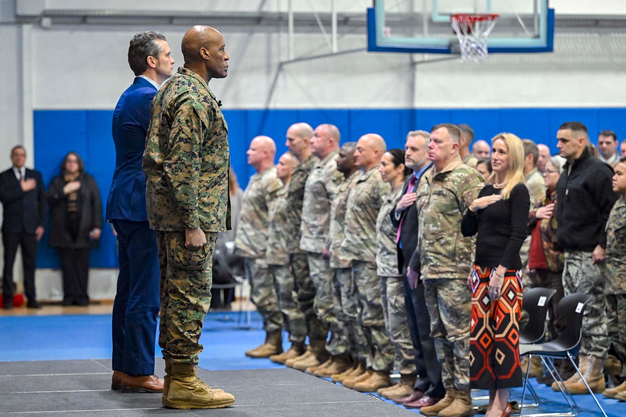 A man in a business suit and a man in a camouflage military uniform are standing on a stage facing a group of uniformed military personnel with their hands on their hearts.