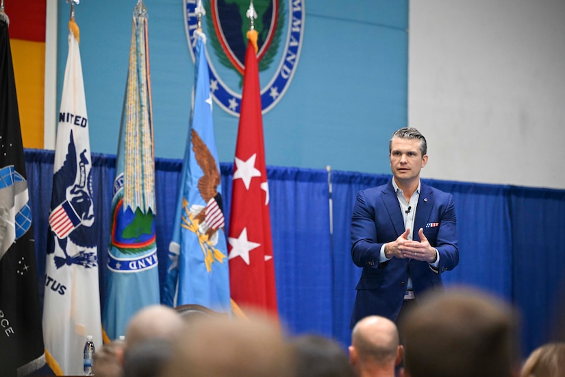 A man in a business suit is talking to seated people with their backs to the camera. There are a number of flags next to the man.