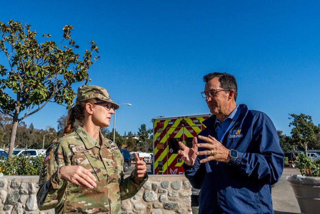 One army general and a civilian chat in a parking lot with an ambulance in the background.