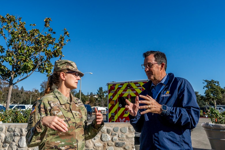 One army general and a civilian chat in a parking lot with an ambulance in the background.