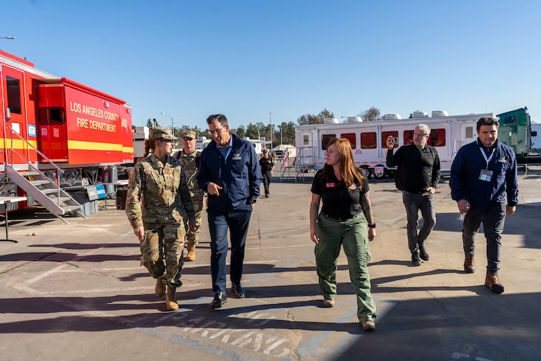 Two Soldiers and four civilians talk and walk in a parking lot. the parking lost has fire department trailers.
