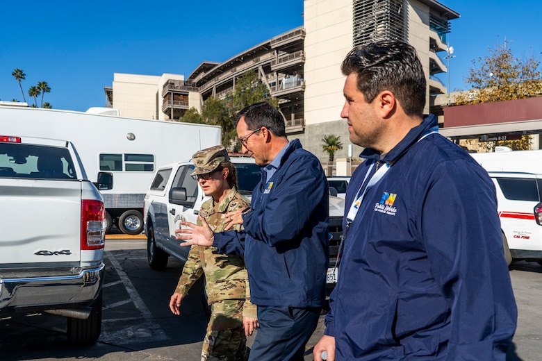 One Army general and two civilians  talk and walk in a parking lot filled with fire department vehicles.