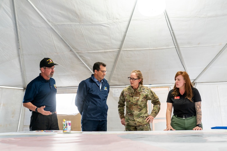 Three civilians and one army general stand a desk in a white tent. they are looking at a large map, which in on the table.