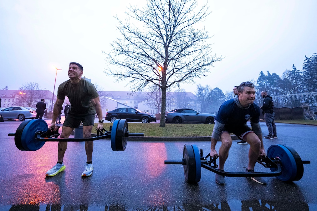 Defense Secretary Pete Hegseth lifts weights outdoors next to a service member, with a tree, buildings and cars in the background.