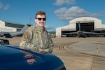 a man leans on the hood of a truck with an aircraft hangar in the background