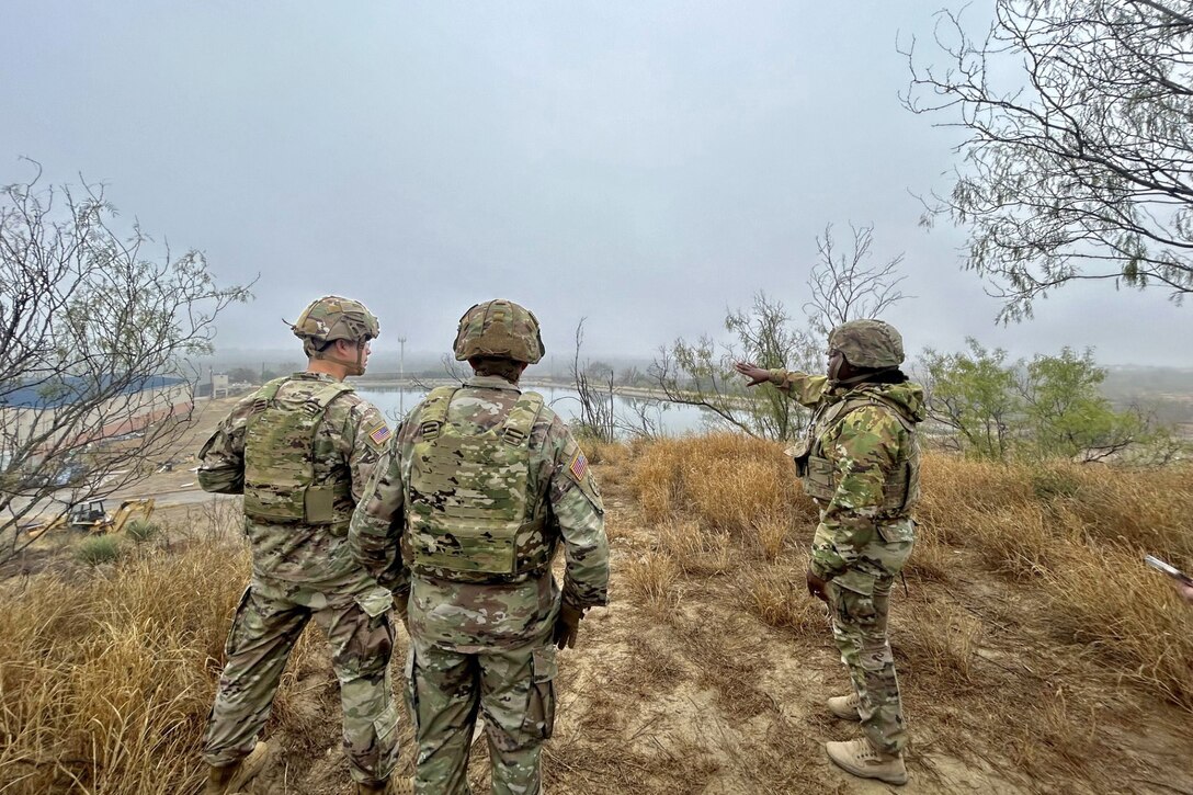 Three soldiers stand in yellow grass while one points to a body of water under overcast skies.