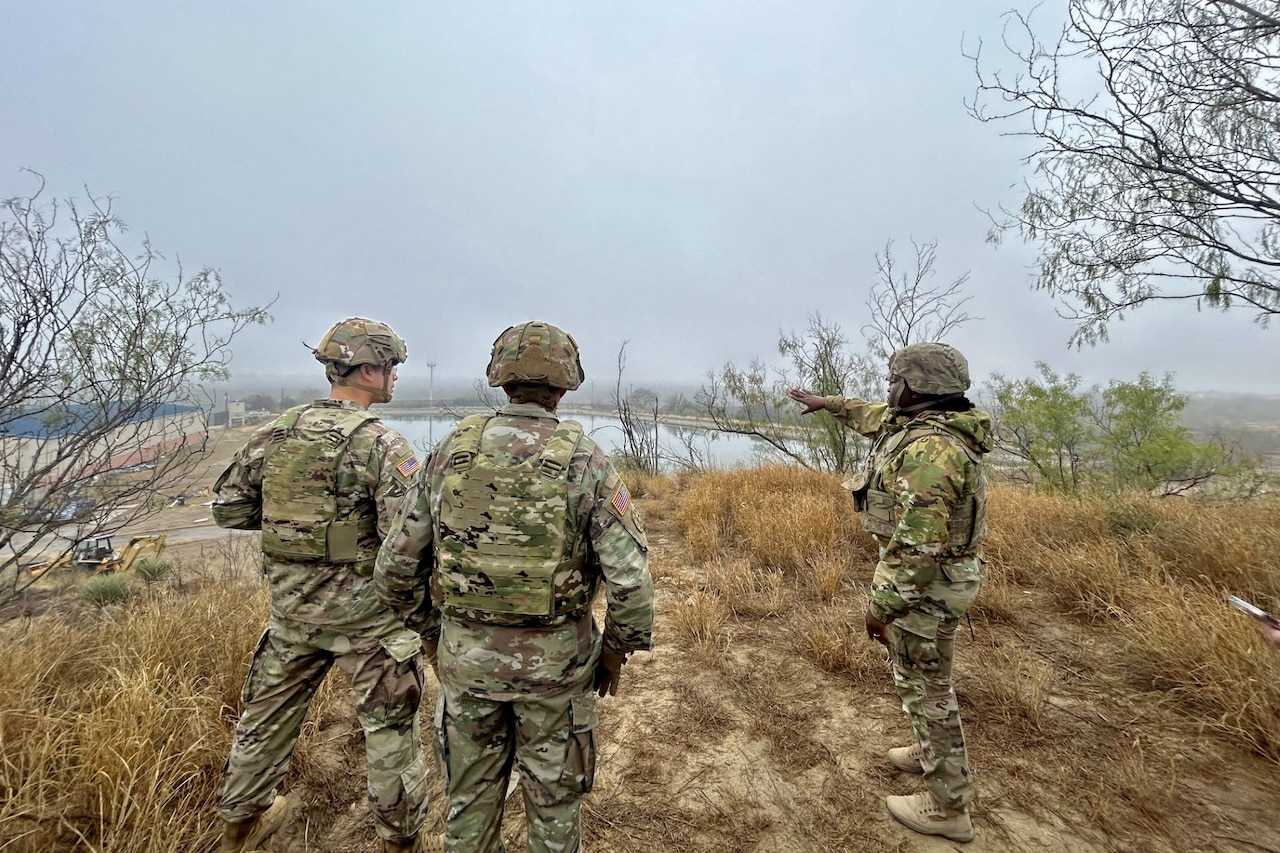 Three soldiers stand in yellow grass while one points to a body of water under overcast skies.