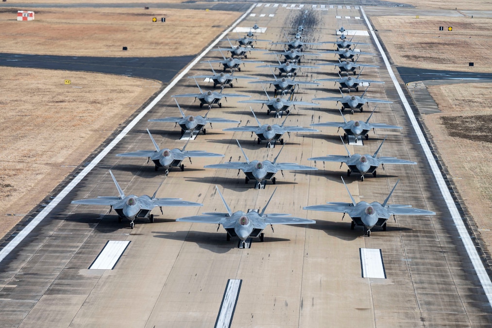 Military aircraft are in rows of three on a flight line during the day.