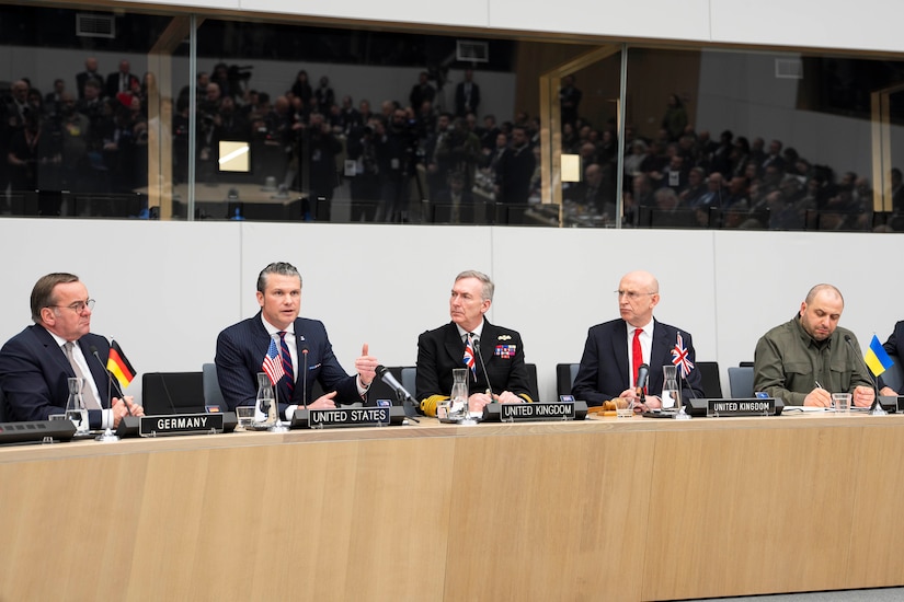 Six men are sitting behind a long table. A man with a placard that reads 