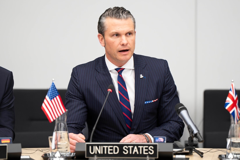 A man in a business suit is sitting behind a microphone at a desk with small U.S. and British flags atop it. There is also a placard on the desk that reads “United States.”