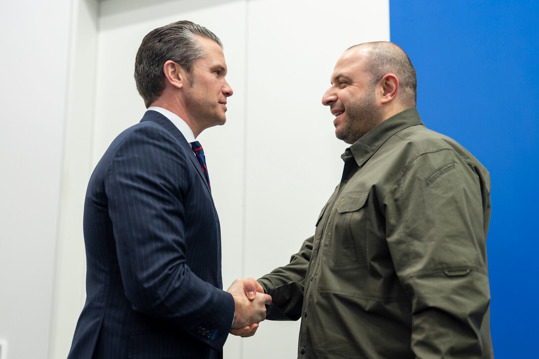 Defense Secretary Pete Hegseth shakes hands with his Ukrainian counterpart in a room with white and blue walls.