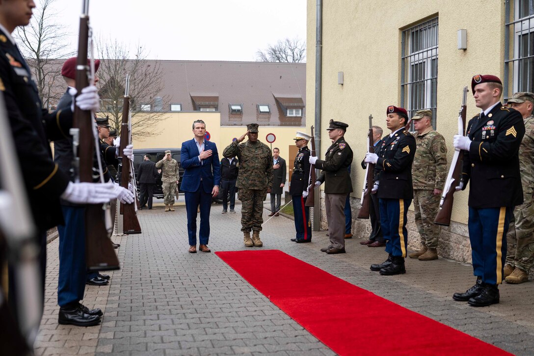 A commander salutes at the end of a red carpet next to a government official holding his hand over his heart.