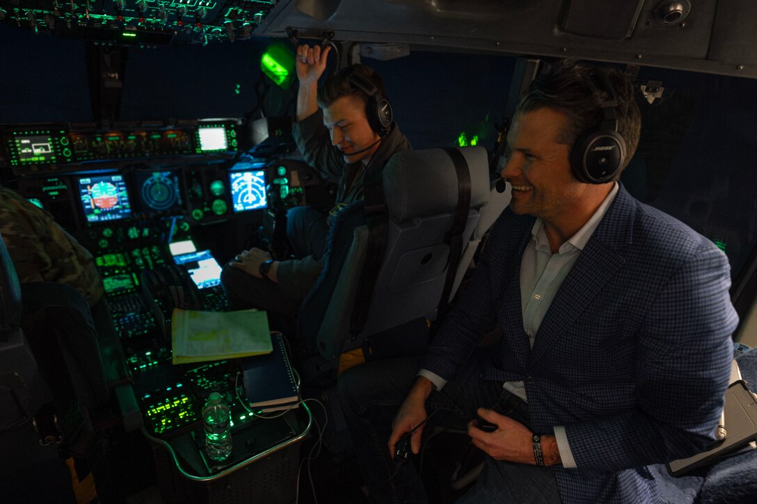 A government official in a suit chats with a pilot and looks at equipment in the cockpit of an aircraft.
