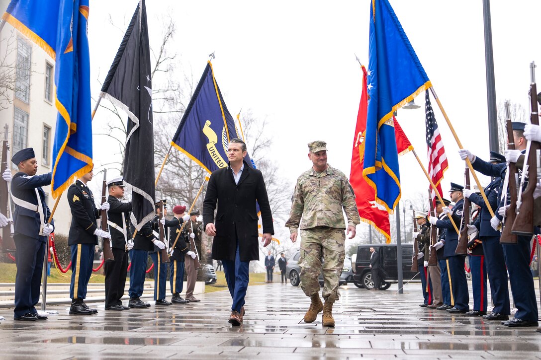 A military officer and a government official walk through an honor guard holding flags during an overcast day.