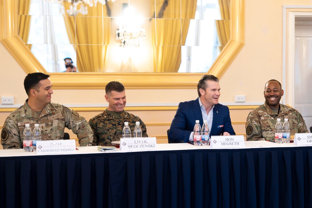 A defense official in a suit sits with soldiers at a table in an elegant room.