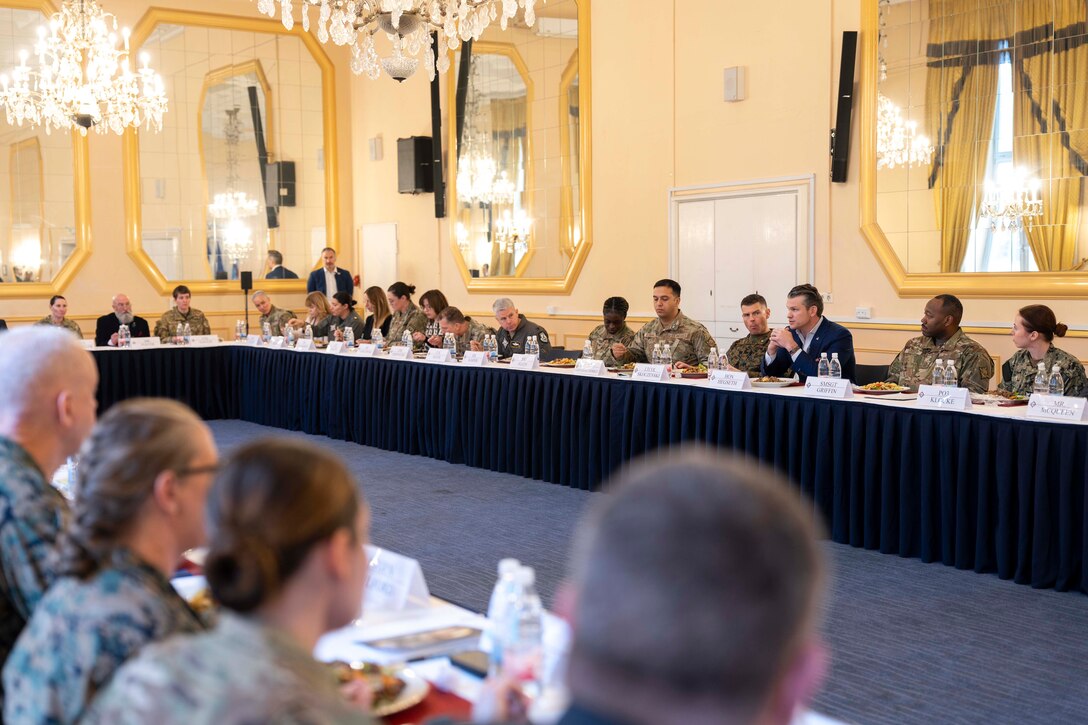 A defense official sits with soldiers in a large table in an elegant room lit by chandelier.