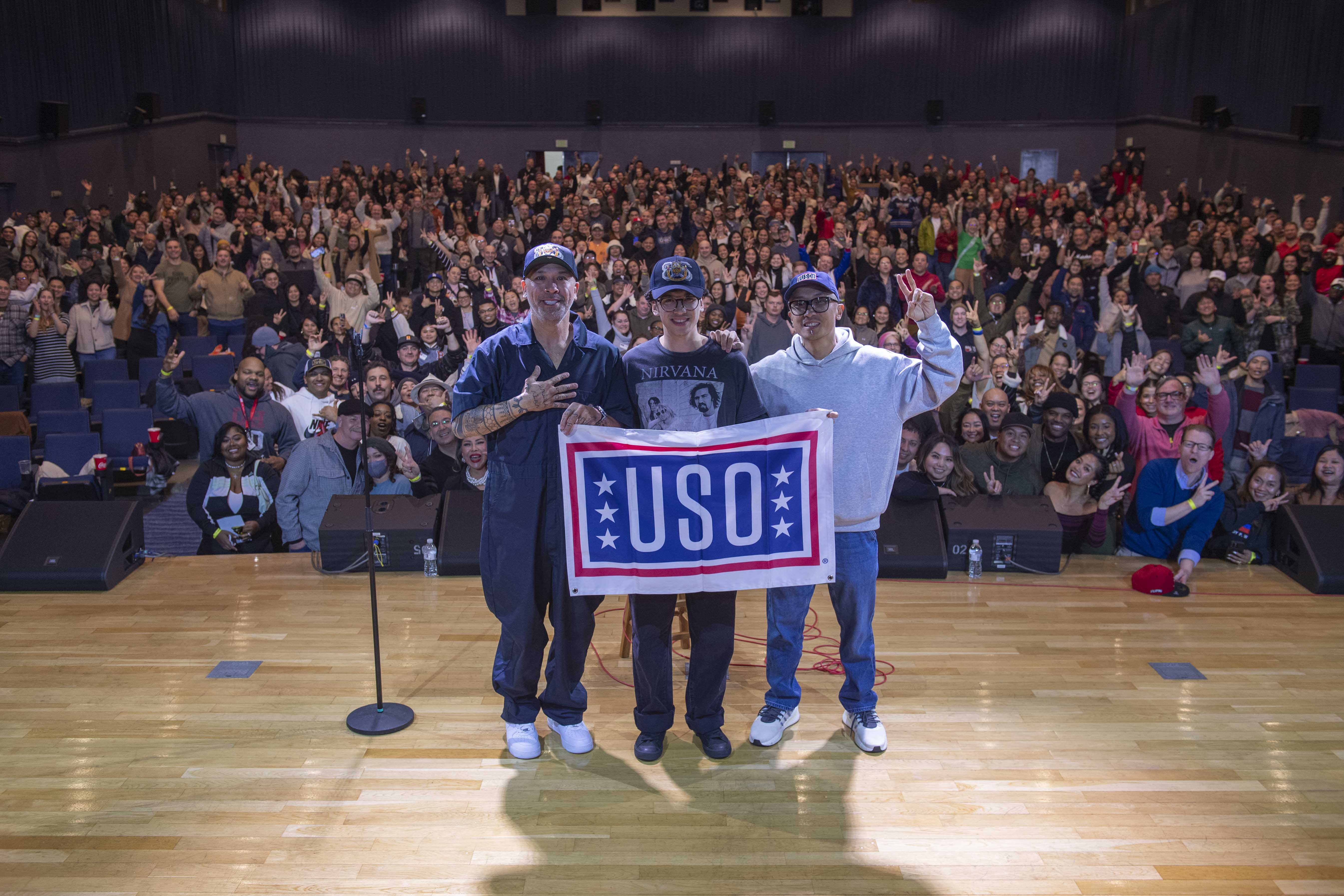 Comedian Jo Koy, his son, and DJ Turbulence pose with a USO banner in front of the audience at the end of the performance at the Morale, Welfare and Recreation (MWR) Fleet Theater at Commander, Fleet Activities Yokosuka.