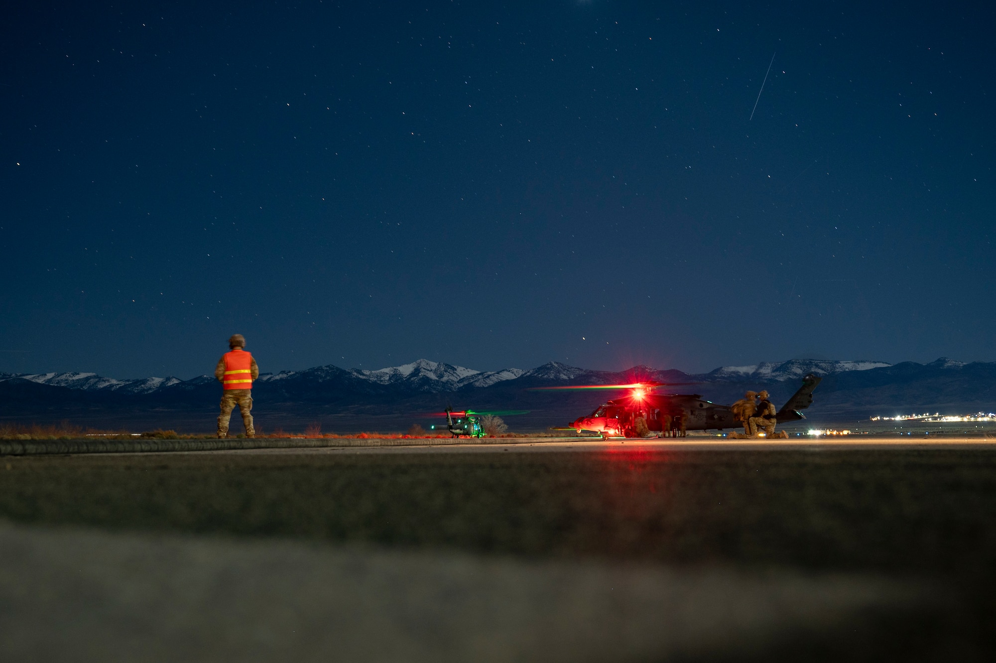 A man facing away from the camera wearing a reflective vest oversees a forward area refueling point exercise. The background features snowcapped mountains and a night sky full of stars.