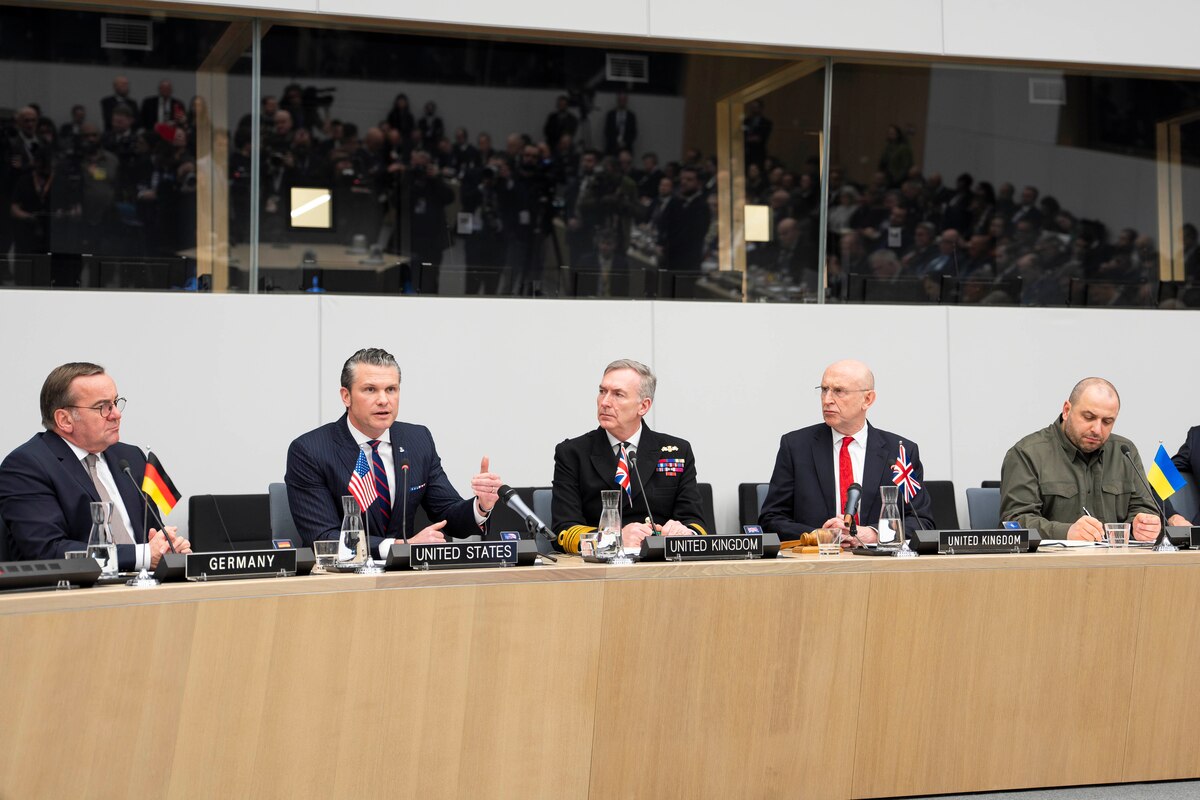 Six men are sitting behind a long table. A man with a placard that reads “United States” is talking.