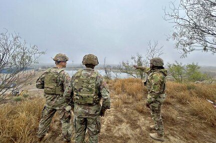 Three soldiers stand in yellow grass while one points to a body of water under overcast skies.