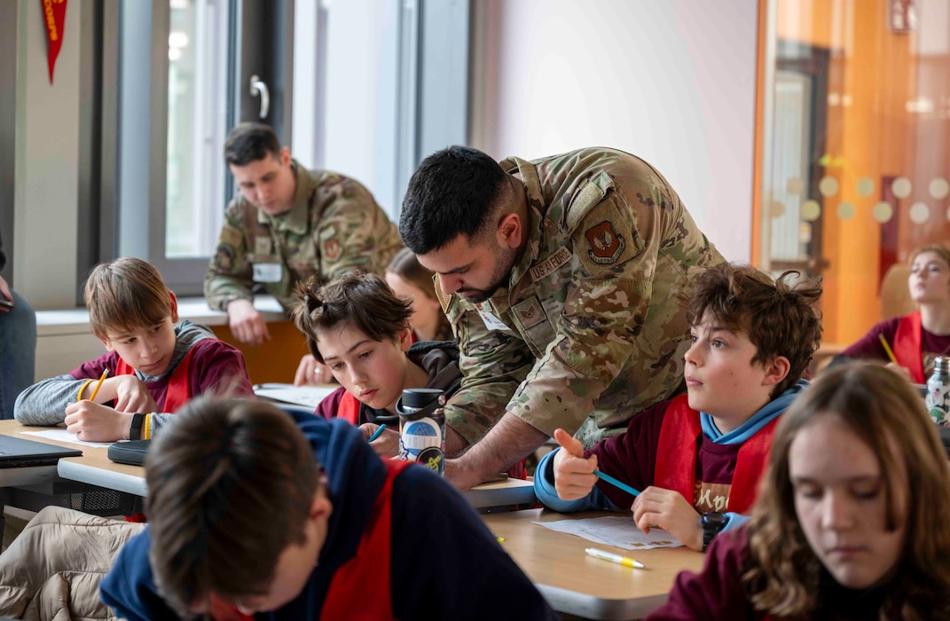 Middle school kids sit at desk next to two Airmen