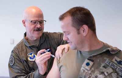 Two men in uniform. One is administering a flu shot to the other.