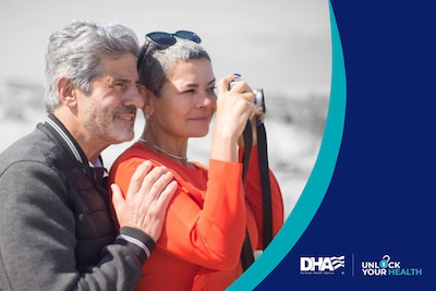 Senior couple (man and woman) with gray hair stand in sweaters on a beach, holding a camera.