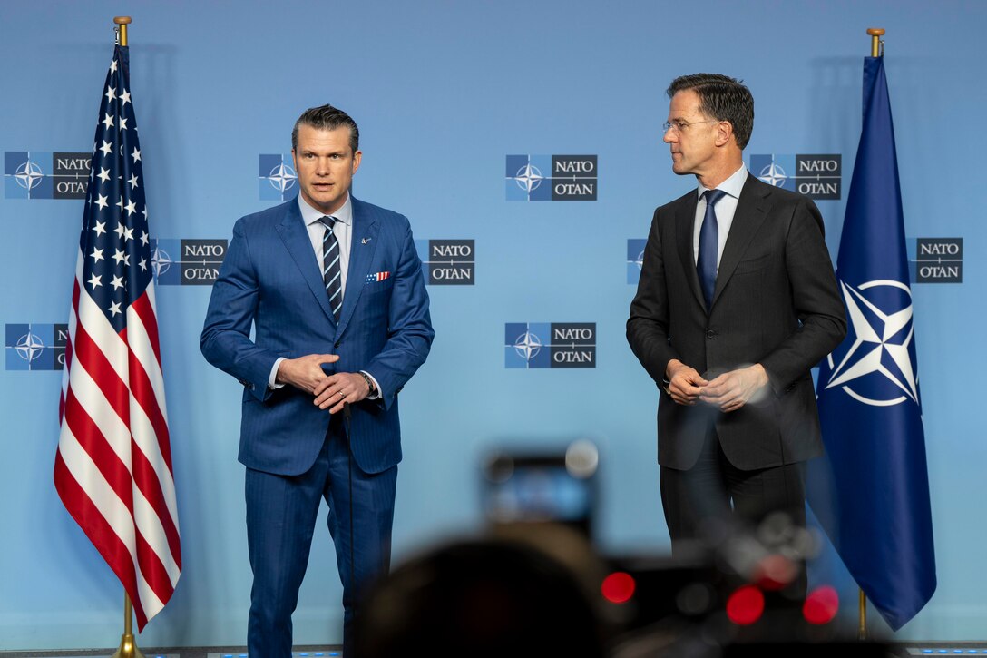 Two men in suits stand in front of an audience in between the U.S. and NATO flags.
