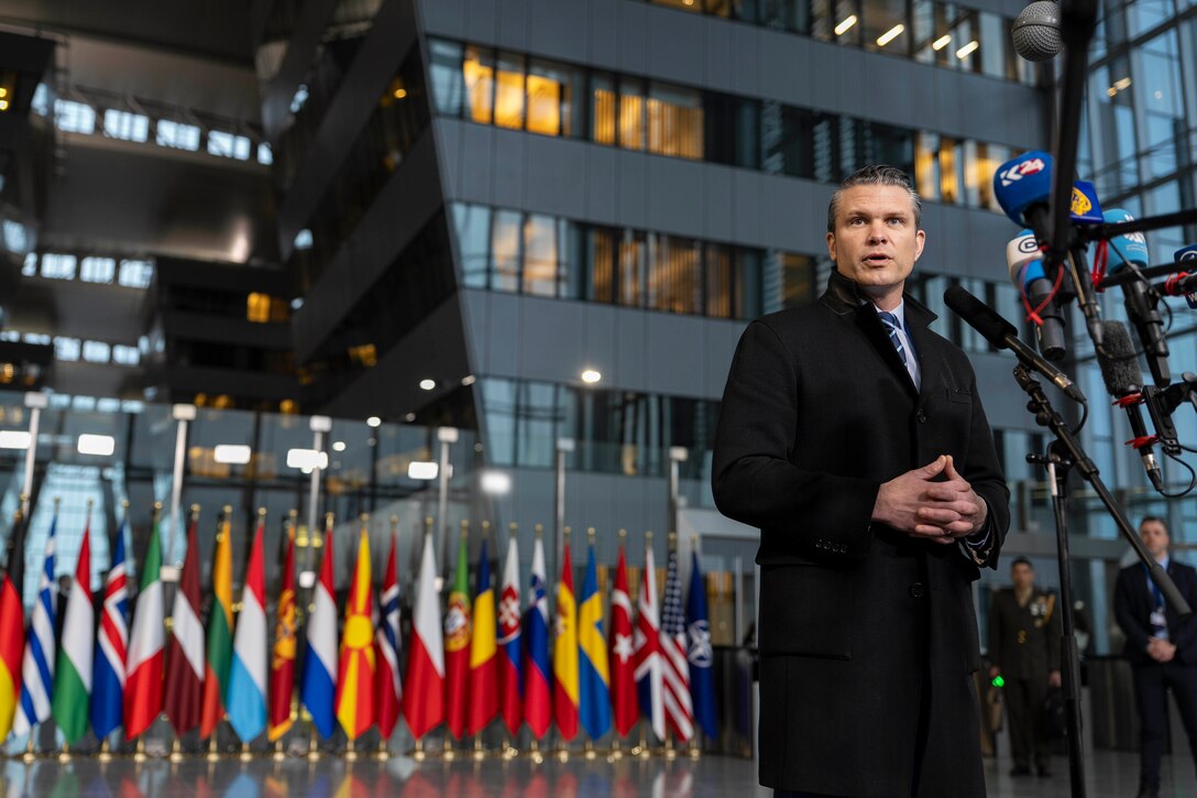 A top government official in a suit jacket speaks into a microphone in front of flags from different countries while in a large lobby.